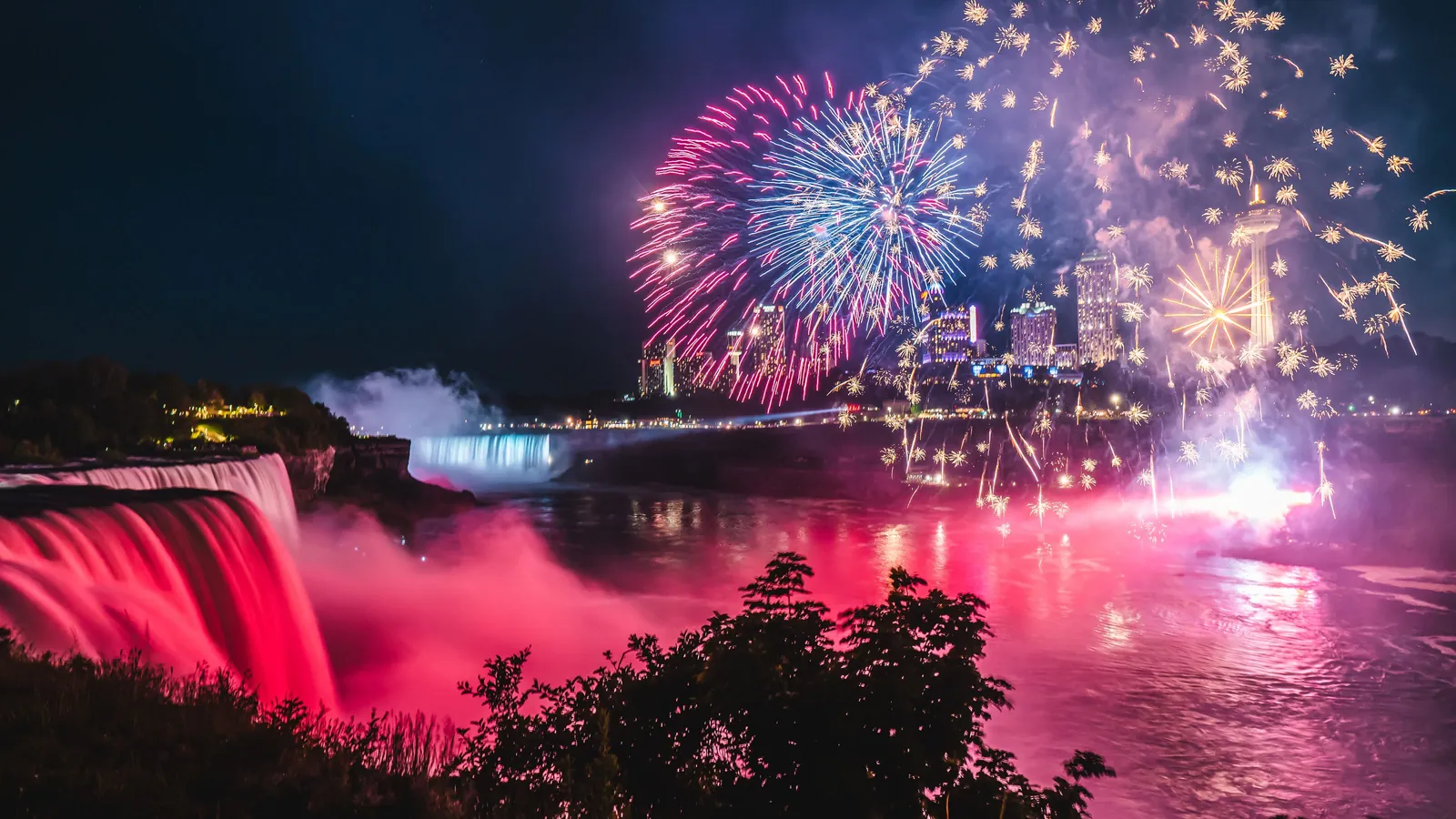 Fireworks bursting over Niagara Falls at night, the water below the falls lit red and the city lights along the horizon
