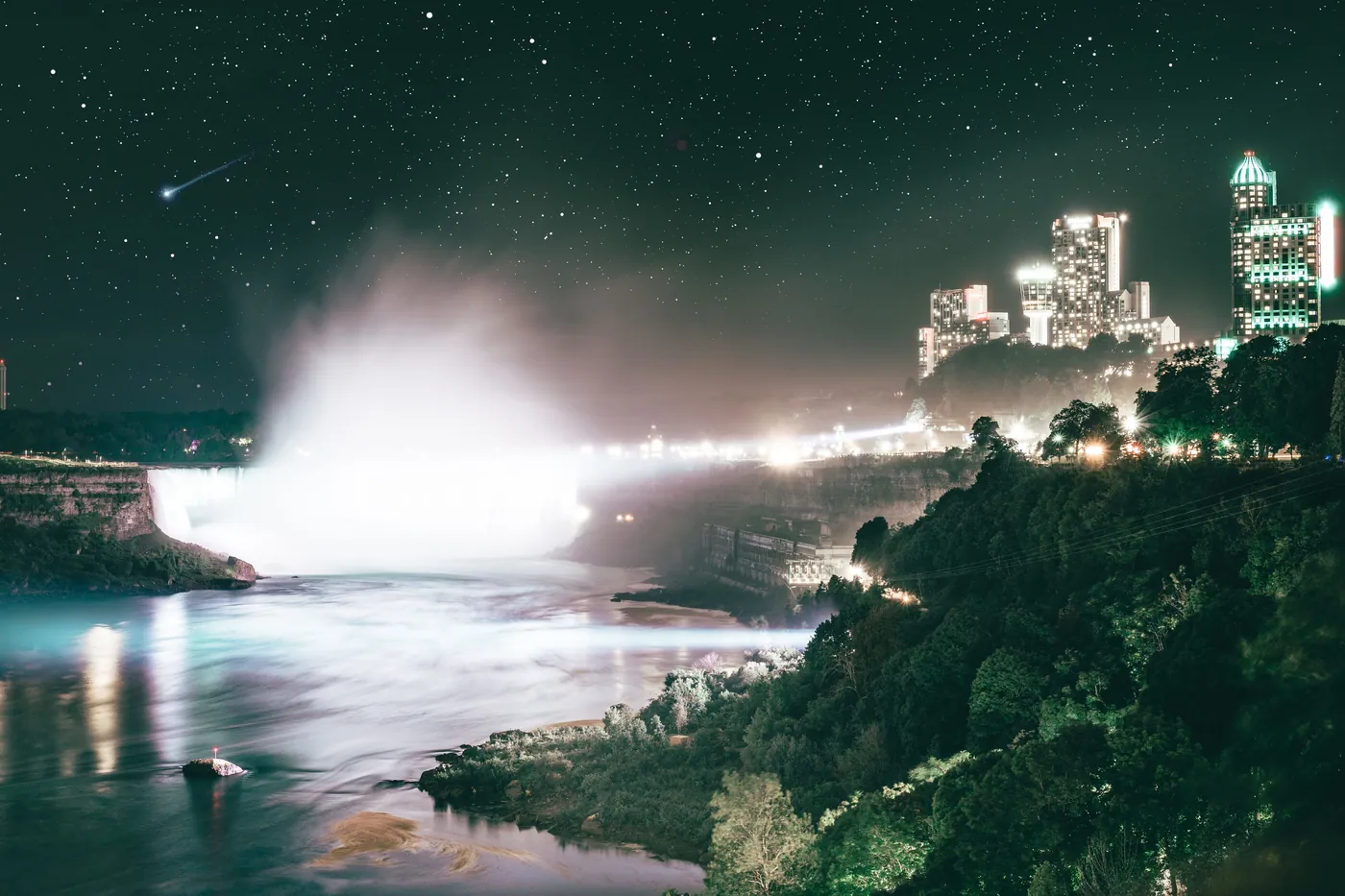 Mist rising from the lit crest of Niagara Falls above the dark gorge, with the lights of the city and a starry sky beyond
