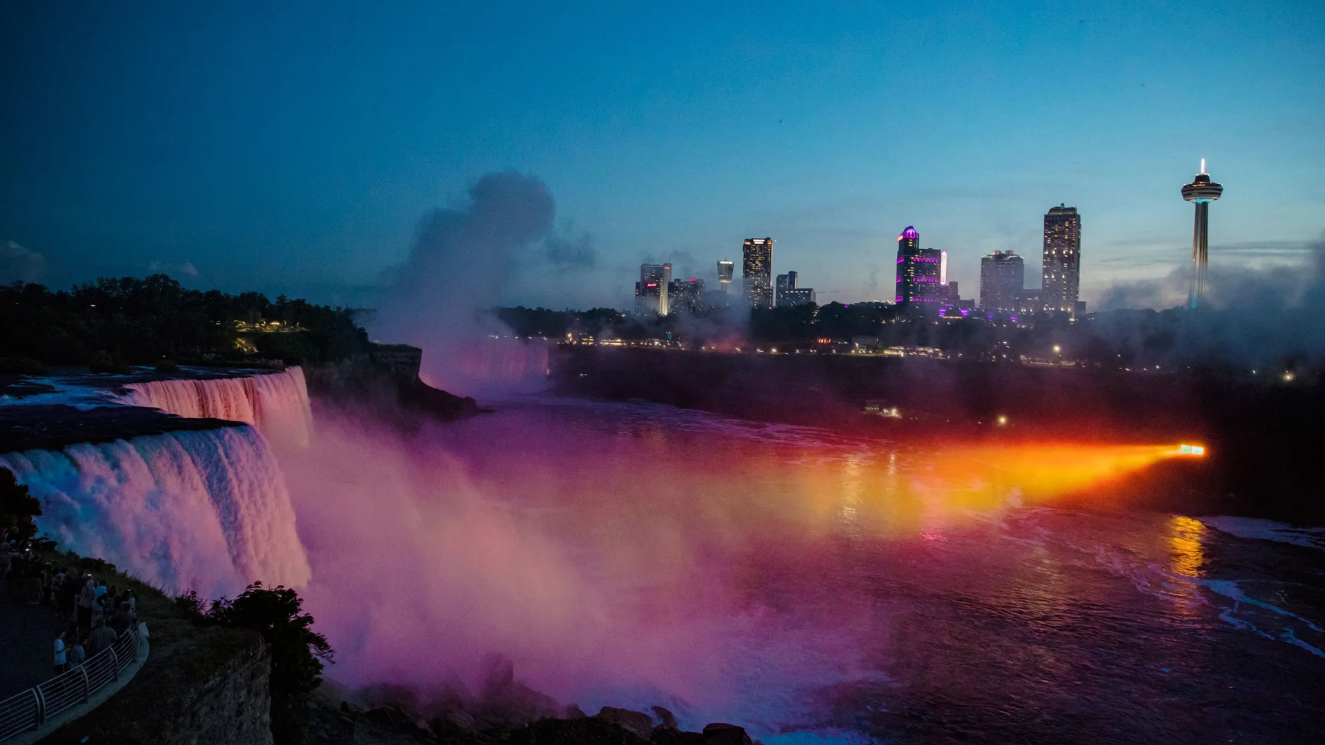 Mist lit pink and violet rising from the foot of Niagara Falls at dusk, with the Skylon Tower and hotel towers of the city beyond and a beam of orange light across the river