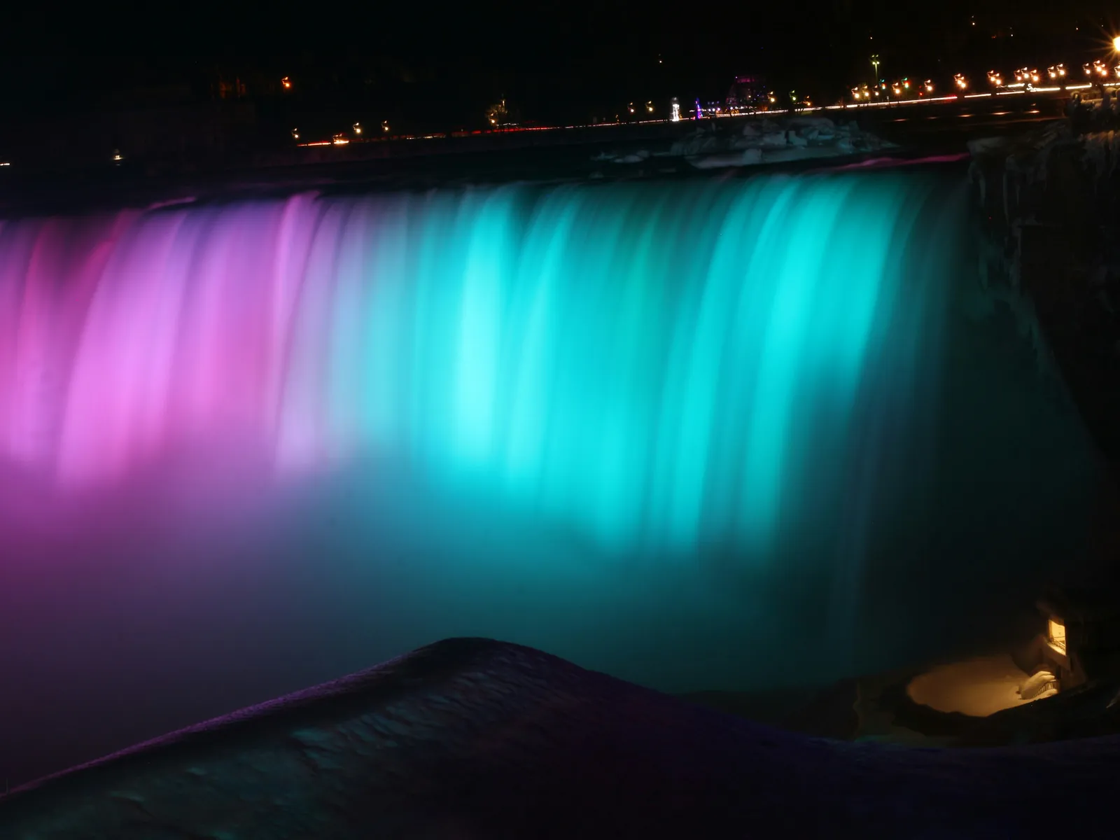 Niagara Falls lit in magenta and teal after dark, seen from the Canadian side, with the lights of the road along the top of the gorge